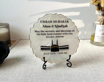 Könnte beinhalten: Eine kreisförmige weiße Harzplatte mit goldfarbenen Rändern, die auf einem schwarzen Metallständer steht. Sie zeigt den Text UMRAH MUBARAK, Adam & Khudijah, einen Segen, eine Grafik der Kaaba und das Datum JULY 2026 / 1448 AH in Schwarz.