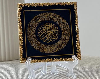 Könnte beinhalten: Eine quadratische schwarze Platte mit komplizierter goldener arabischer Kalligrafie, die in einem kreisförmigen Rand zentriert ist. Die Kanten der Platte sind mit strukturierten Blattgoldfragmenten verziert. Das Stück steht auf einem transparenten Kunststoffständer.