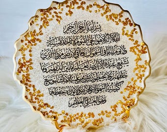 Könnte beinhalten: Eine kreisförmige Harzplatte mit gewelltem Rand, die eine weiße und goldglitzernde Basis mit eingebetteten Blattgoldflocken aufweist. In der Mitte befindet sich die schwarze arabische Kalligrafie des Thronverses. Die Platte steht auf einem kleinen goldfarbenen Metallständer.