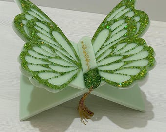 Könnte beinhalten: Ein hellgrüner, schmetterlingsförmiger Buchständer mit Glitzerakzenten. Die Flügel sind mit Adern und Glitzer versehen, und die Mitte hat goldenen Text. Eine goldene Quaste hängt von unten.