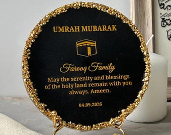 Könnte beinhalten: Eine kreisförmige schwarze Plakette mit goldenem, strukturiertem Rand auf einem goldenen Metallständer. Sie zeigt den Text UMRAH MUBARAK, ein Kaaba-Symbol, Farooq Family und den Text May the serenity and blessings of the holy land remain with you always. Ameen. 04.09.2026.
