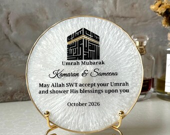 Könnte beinhalten: Eine kreisförmige weiße Harzplatte mit goldenem Rand auf einem goldenen Metallständer. Sie zeigt eine schwarze Kaaba-Grafik sowie den Text "Umrah Mubarak", "Kamaran & Sameena", "May Allah SWT accept your Umrah and shower His blessings upon you" und "October 2026".