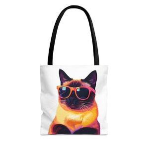 Puede incluir: Un bolso tote blanco con asas negras que presenta un gráfico colorido de un gato siamés con gafas de sol de color naranja brillante. El gato tiene marcas faciales de color marrón oscuro y un pecho color crema, en una ilustración estilizada.