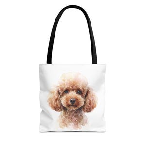 Puede incluir: Un bolso tote blanco con una ilustración en acuarela de un caniche marrón. El perro está centrado en el bolso, mostrando pinceladas suaves y difuminadas en tonos beige y marrón claro. El bolso incluye dos asas de tela de color negro.