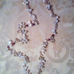 May include: A white pearl necklace with burgundy beads and matching earrings. The necklace is made of irregular shaped pearls and is strung on a gold chain.