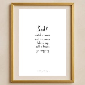 Può includere: Una stampa incorniciata con una cornice color oro. La stampa presenta la parola "Sad?" in alto, seguita da suggerimenti: guarda un film, mangia un gelato, fai un pisolino, chiama un amico e vai a fare shopping. Il testo è in stile scritto a mano.