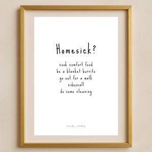 Può includere: Una stampa incorniciata con una cornice color oro. La carta bianca all'interno ha la parola "Homesick?" in alto, seguita da un elenco di attività confortanti in testo nero. La parte inferiore della stampa ha le parole "mandy_reminding".