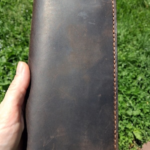 May include: A rectangular dark brown leather wallet featuring a smooth, matte finish with visible natural grain variations. The edges are secured with thick, contrasting tan stitching that runs along the entire perimeter of the wallet's front face.