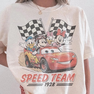 K&ouml;nnte beinhalten: Elfenbeinfarbenes T-Shirt mit einem Aufdruck von Mickey Mouse, Donald Duck, Daisy Duck und Minnie Mouse in einem roten Rennwagen mit dem Text "Speed Team 1928". Zwei karierte Flaggen im Hintergrund.