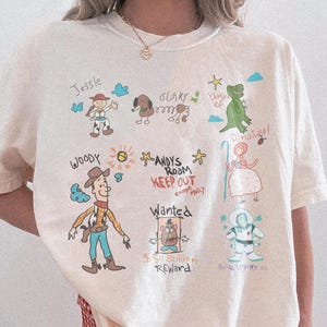 Puede incluir: Camiseta Comfort Colors color marfil con dibujos de personajes de dibujos animados como Woody y Jessie. La camiseta incluye el texto "ANDY'S ROOM KEEP OUT" y "Wanted".
