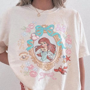 Puede incluir: Camiseta de color marfil con una ilustraci&oacute;n enmarcada de una princesa pelirroja, rodeada de elementos florales y acu&aacute;ticos. El dise&ntilde;o incluye una perla, estrellas y un cangrejo rojo. El texto "Comfort Colors Ivory" est&aacute; impreso en la parte inferior derecha.