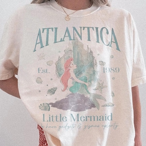 Może przedstawiać: Kremowa koszulka z nadrukiem z napisem "ATLANTICA" i "Little Mermaid". Projekt zawiera ilustrację syreny, zamek i tekst "Est. 1989". Koszulka jest częścią kolekcji Comfort Colors.