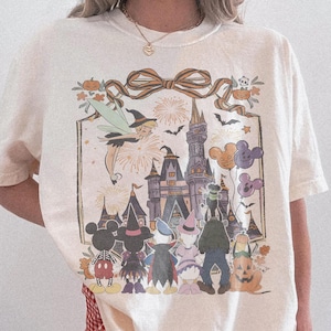 Pode incluir: Camiseta Comfort Colors marfim com um gr&aacute;fico de Halloween. O design inclui um castelo, personagens, fogos de artif&iacute;cio e uma fada. A camisa tem um ajuste folgado e mangas curtas. O texto "Comfort Colors Ivory" &eacute; impresso na parte inferior direita.