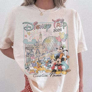 May include: Cream-coloured t-shirt with a Disney Trip 2026 graphic featuring Mickey Mouse and friends. The design includes a castle and fireworks. The shirt has a crew neck and short sleeves. The words "Custom Name" are printed below the graphic.