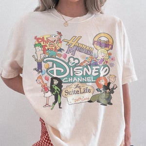 Peut inclure: T-shirt ivoire avec un motif Disney Channel représentant des personnages de séries comme Phineas et Ferb, Hannah Montana et Kim Possible. Le t-shirt a un col rond et des manches courtes. Le texte "Comfort Colors Ivory" est imprimé en bas à droite.
