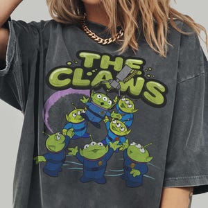 May include: Dark gray t-shirt featuring a graphic of green aliens in blue uniforms with the text "THE CLAWS". The design includes a purple arc and a claw machine. The shirt is from Comfort Colors.