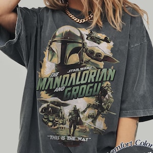 Może przedstawiać: Ciemnoszara koszulka z grafiką z Gwiezdnych Wojen: The Mandalorian. Projekt zawiera hełm Mandalorianina, Grogu i tekst "THE MANDALORIAN AND GROGU". Na koszulce wydrukowano r&oacute;wnież frazę "THIS IS THE WAY".