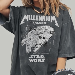 Può includere: T-shirt grigio scuro con una grafica dell'astronave Millennium Falcon, la scritta "MILLENNIUM FALCON" e "STAR WARS". La maglietta ha anche la scritta "Ride the lightning". La maglietta è un Comfort Colors Pepper.