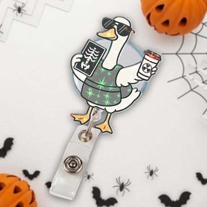 May include: A Halloween-themed badge reel featuring a cartoon goose wearing sunglasses and a medical vest, holding an X-ray and a coffee cup with a radiation symbol. The badge reel has a clear plastic clip and a retractable cord.