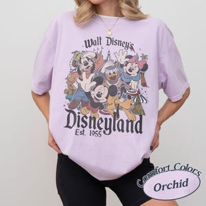 May include: A light purple t-shirt featuring a vintage Disneyland graphic with Mickey Mouse, Minnie Mouse, Donald Duck, Goofy, and Pluto. The shirt has short sleeves and the text "Walt Disney's Disneyland Est. 1955".