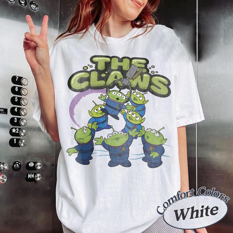 May include: White t-shirt featuring a graphic of green aliens in blue uniforms with the text "THE CLAWS". The design includes a purple ring and a claw machine. The shirt is labeled "Comfort Colors White".