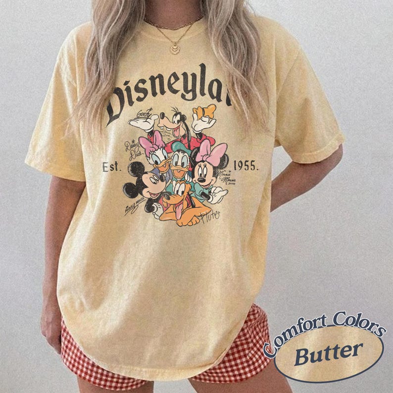 May include: A light yellow t-shirt featuring the word "Disneyland" in a vintage font, along with a graphic of Disney characters. The shirt includes the text "Est. 1955." and the Comfort Colors Butter logo.