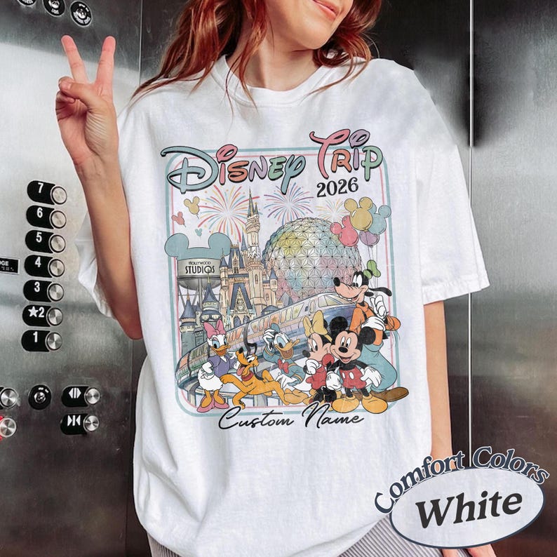 May include: White t-shirt with a Disney Trip 2026 design, including Mickey Mouse, Donald Duck, and Goofy. The shirt features the text "Custom Name" and "Comfort Colors White". The design includes fireworks and a castle.