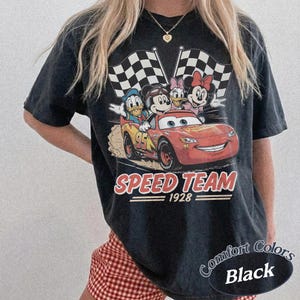K&ouml;nnte beinhalten: Schwarzes T-Shirt mit einer Grafik von Mickey Mouse, Donald Duck, Daisy Duck und Minnie Mouse in einem roten Rennwagen. Der Text "SPEED TEAM 1928" steht darunter, mit zwei karierten Flaggen.