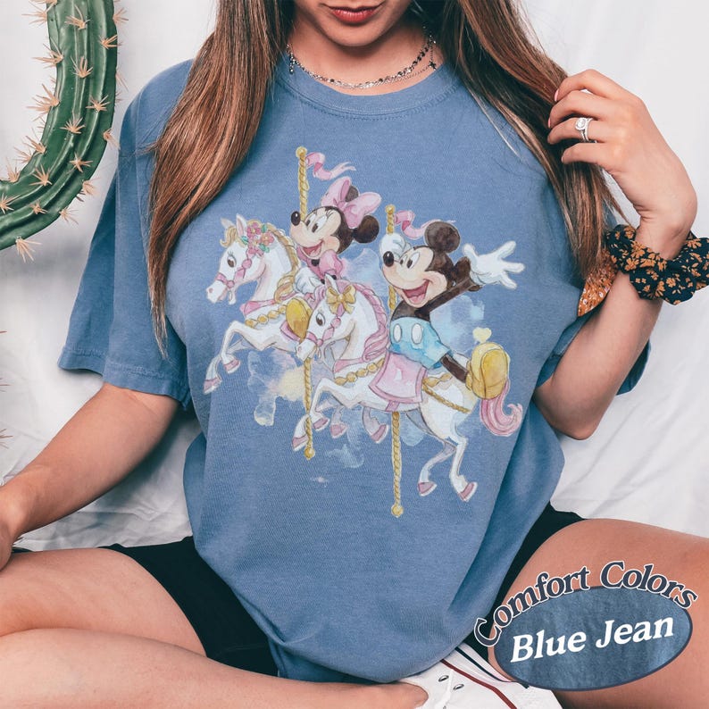 Puede incluir: Camiseta Comfort Colors azul vaquero con un dise&ntilde;o de carrusel con Mickey Mouse y Minnie Mouse montados en caballos blancos. El gr&aacute;fico incluye detalles en rosa y amarillo. La camiseta tiene mangas cortas.