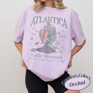 Może przedstawiać: Jasnofioletowa koszulka z grafiką syreny i napisami "ATLANTICA" i "Little Mermaid". Koszulka zawiera r&oacute;wnież tekst "Est. 1989" i "We have gadgets & gizmos aplenty". Koszulka jest oznaczona jako "Comfort Colors Orchid".