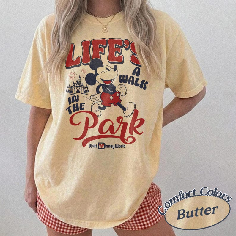 May include: A light yellow t-shirt with a graphic featuring Mickey Mouse and the text "LIFE'S A WALK IN THE PARK" in red and black. The shirt also includes the words "Walt Disney World". The shirt is a Comfort Colors Butter.