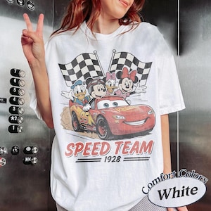 K&ouml;nnte beinhalten: Wei&szlig;es T-Shirt mit einem Cartoon-Motiv von Micky Maus, Donald Duck, Daisy Duck und Minnie Maus in einem roten Rennwagen mit karierten Flaggen. Der Text "SPEED TEAM 1928" ist darunter gedruckt.