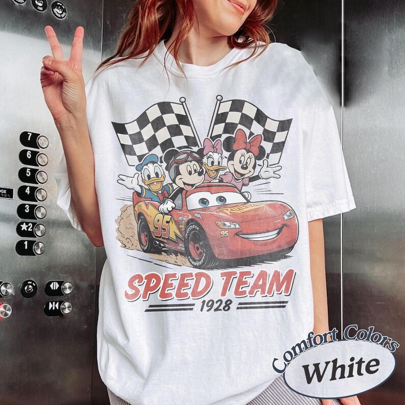 K&ouml;nnte beinhalten: Wei&szlig;es T-Shirt mit einem Cartoon-Motiv von Micky Maus, Donald Duck, Daisy Duck und Minnie Maus in einem roten Rennwagen mit karierten Flaggen. Der Text "SPEED TEAM 1928" ist darunter gedruckt.