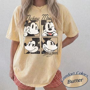 Op de afbeelding: Lichtgeel T-shirt met een vierdelige Mickey Mouse-afbeelding. Het shirt toont de tekst "Mickey Mouse" boven de afbeelding en "Comfort Colors Butter" in een cirkelvormig ontwerp. Een casual kledingstuk.