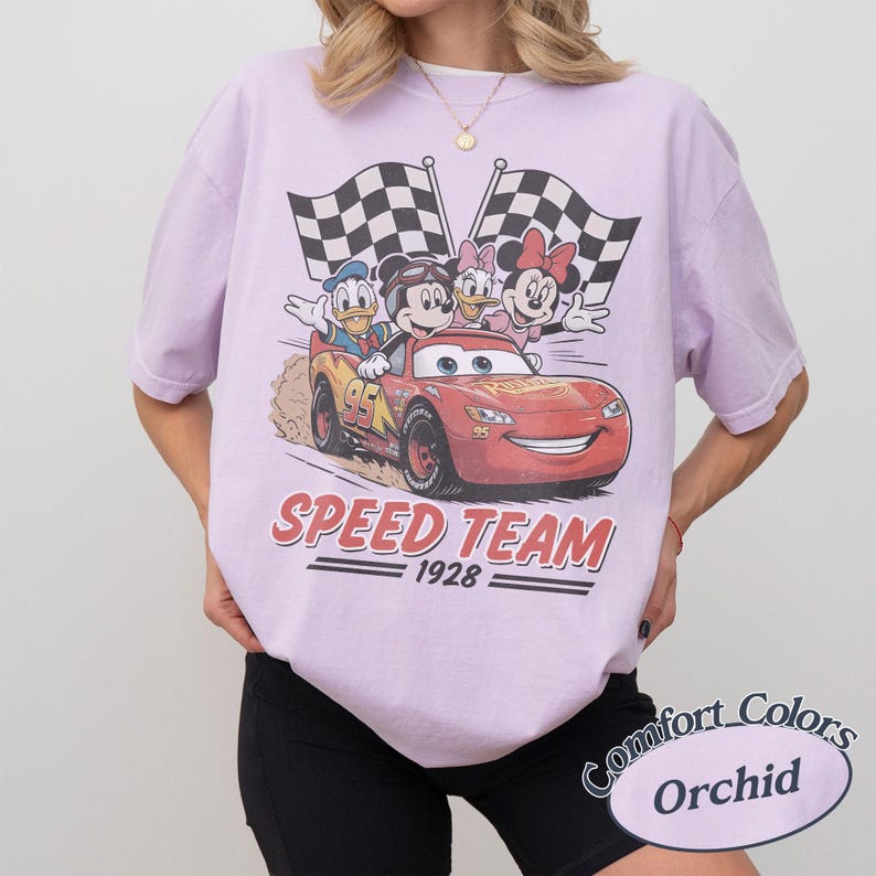 K&ouml;nnte beinhalten: Ein helllila T-Shirt mit einem Aufdruck von Disney-Figuren in einem roten Rennwagen. Das Design zeigt Micky Maus, Donald Duck, Daisy Duck und Minnie Maus mit karierten Flaggen. Der Text "SPEED TEAM 1928" ist ebenfalls zu sehen.