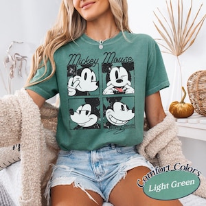 Op de afbeelding: Lichtgroen T-shirt met een vierpaneelsafbeelding van Mickey Mouse met verschillende uitdrukkingen. De woorden "Mickey Mouse" staan boven de afbeelding. Het shirt is gelabeld met "Comfort Colors" en "Light Green".