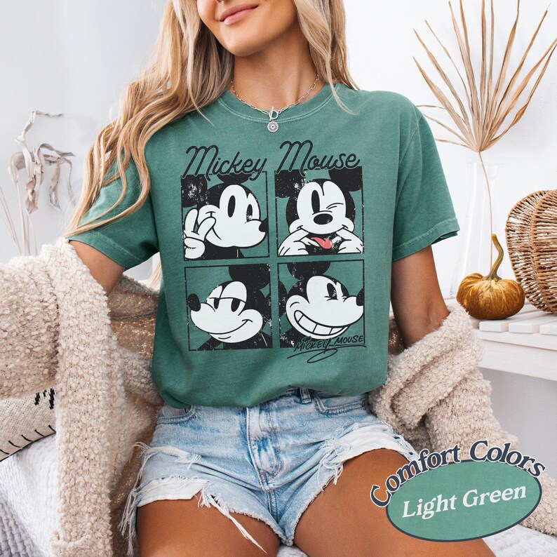 Op de afbeelding: Lichtgroen T-shirt met een vierpaneelsafbeelding van Mickey Mouse met verschillende uitdrukkingen. De woorden "Mickey Mouse" staan boven de afbeelding. Het shirt is gelabeld met "Comfort Colors" en "Light Green".