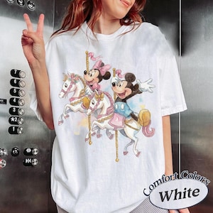 Puede incluir: Camiseta blanca con un dise&ntilde;o de carrusel con Mickey y Minnie Mouse montados en caballos blancos. El dise&ntilde;o incluye detalles en rosa y amarillo. La camiseta tiene el texto "Comfort Colors White" en un dise&ntilde;o circular.