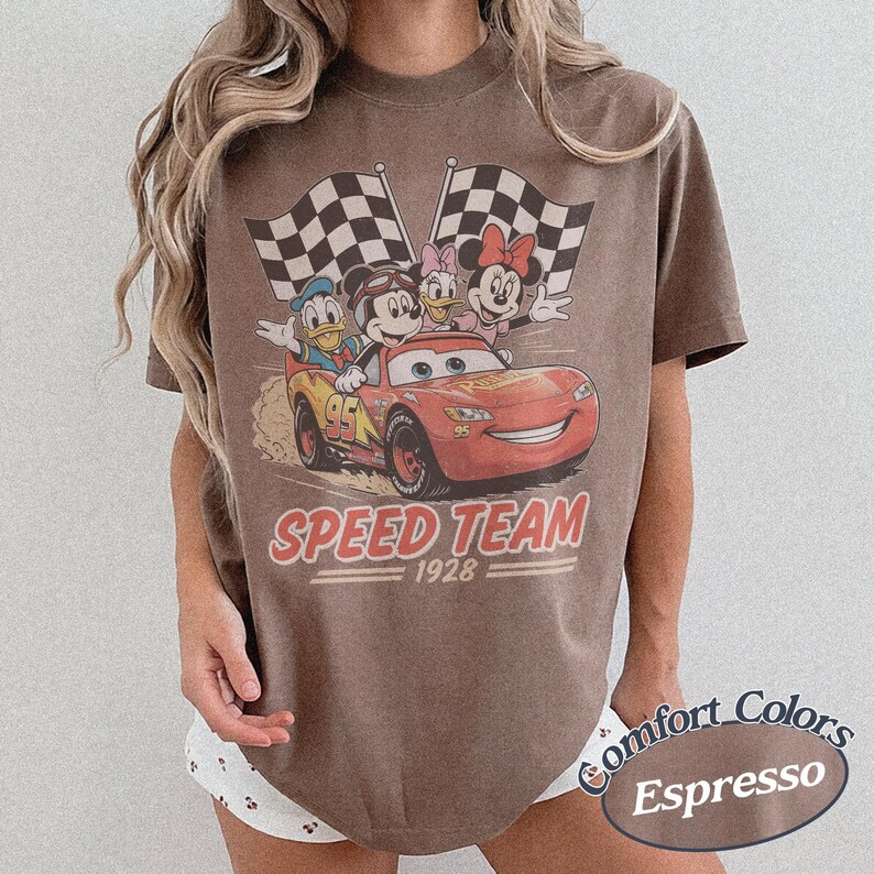 K&ouml;nnte beinhalten: Espressofarbenes T-Shirt mit einem Retro-Grafikdesign von Disney-Figuren in einem Rennwagen. Das Design zeigt Micky Maus, Donald Duck, Daisy Duck und Minnie Maus mit dem Text "SPEED TEAM 1928".