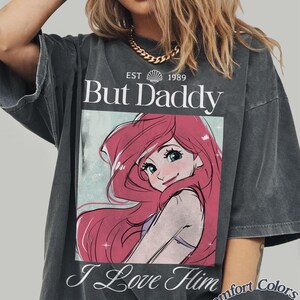 K&ouml;nnte beinhalten: Dunkelgraues T-Shirt mit dem Text "But Daddy I Love Him" und einer Cartoon-Grafik mit roten Haaren und blauen Augen. Das Shirt ist von Comfort Colors.
