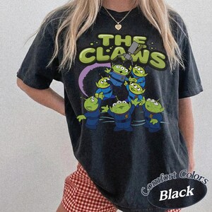 May include: Black t-shirt featuring a graphic of green aliens in blue uniforms with the text "THE CLAWS". The design includes a purple arc and a claw machine. The shirt is labeled "Comfort Colors Black".