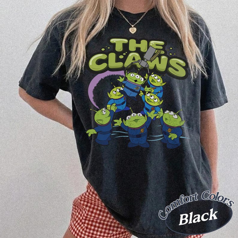 May include: Black t-shirt featuring a graphic of green aliens in blue uniforms with the text "THE CLAWS". The design includes a purple arc and a claw machine. The shirt is labeled "Comfort Colors Black".