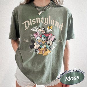 May include: Moss green t-shirt featuring the Disneyland logo and a graphic of Mickey Mouse, Minnie Mouse, Donald Duck, Daisy Duck, Goofy, and Pluto. The shirt includes the text "Est. 1955" and "Comfort Colors Moss".