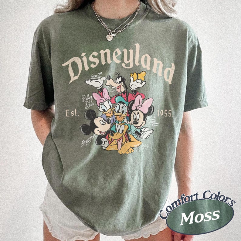 May include: Moss green t-shirt featuring the Disneyland logo and a graphic of Mickey Mouse, Minnie Mouse, Donald Duck, Daisy Duck, Goofy, and Pluto. The shirt includes the text "Est. 1955" and "Comfort Colors Moss".