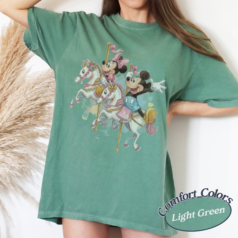 Puede incluir: Camiseta Comfort Colors verde claro con una escena de carrusel con Mickey y Minnie Mouse montados en caballos blancos. La camiseta tiene un corte holgado y las palabras "Comfort Colors Light Green" est&aacute;n impresas en la parte inferior derecha.
