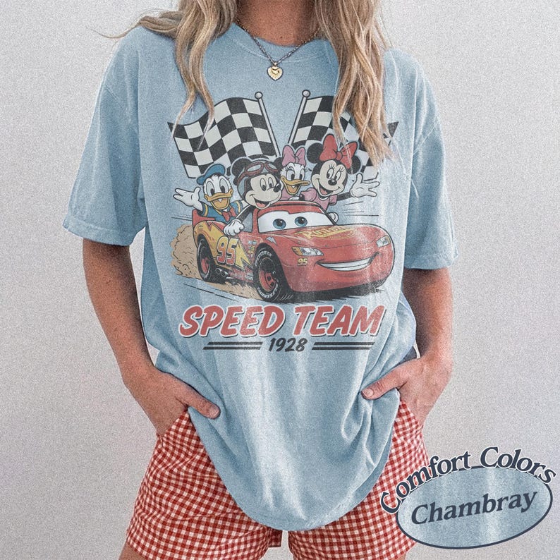 K&ouml;nnte beinhalten: Hellblaues T-Shirt mit einem Vintage-Grafikdesign von Disney-Figuren in einem Rennwagen. Das Design enth&auml;lt den Text "SPEED TEAM 1928" und Schachflaggen. Das T-Shirt hat eine lockere Passform.