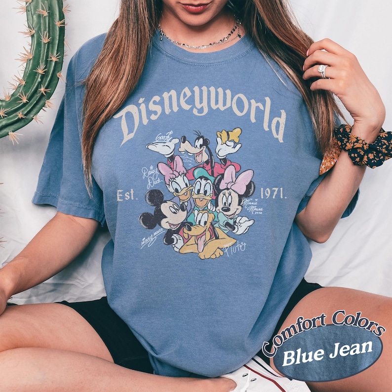 Może przedstawiać: Niebieska koszulka z grafiką Disney World przedstawiającą Myszka Miki, Minnie, Kaczora Donalda, Daisy, Goofiego i Plutona. Koszulka zawiera napis "Disneyworld Est. 1971" i "Comfort Colors Blue Jean".