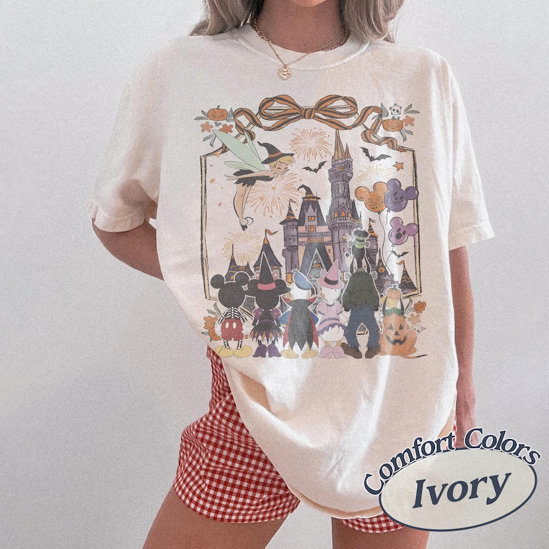 Pode incluir: Camiseta Comfort Colors marfim com um gr&aacute;fico de Halloween. O design inclui um castelo, personagens, fogos de artif&iacute;cio e uma fada. A camisa tem um ajuste folgado e mangas curtas. O texto "Comfort Colors Ivory" &eacute; impresso na parte inferior direita.