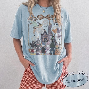 Pode incluir: Camiseta azul claro com um gr&aacute;fico com um castelo, personagens e fogos de artif&iacute;cio. O design inclui uma fada, morcegos e bal&otilde;es. A camisa tem as palavras "Comfort Colors Chambray" impressas.