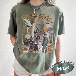 Pode incluir: T-shirt verde musgo com um gr&aacute;fico de Halloween com um castelo, personagens e ab&oacute;boras. O design inclui uma bruxa, fogos de artif&iacute;cio e bal&otilde;es em forma de Mickey Mouse. A camisa tem uma etiqueta Comfort Colors.